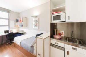 A typical residence hall in Madrid furnished with a desk and bookshelf, a twin sized bed. Room includes a kitchenette which includes a microwave, small refrigerator, and sink. 